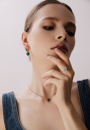 Bijoux en ton doré mettant en valeur des pierres précieuses vertes en forme de cœur. Le collier dispose d'une chaîne ; les boucles d'oreilles et une bague complètent le design.