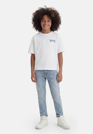 Enfant aux cheveux bouclés portant un t-shirt blanc « Replay », un jean bleu clair et des baskets blanches montantes, debout et souriant.