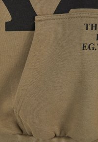 Felpa di colore beige con una texture morbida. Presenta una stampa grafica nera e una tasca inclinata. Polsini a costine sul fondo.