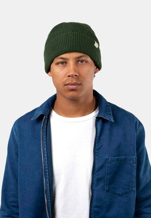 Green knitted beanie with a ribbed texture; worn by a person with a blue jacket over a white shirt. Simple design, no visible patterns.