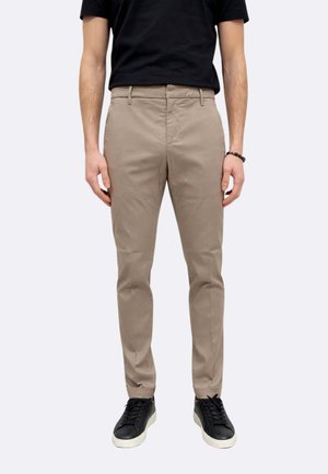 Uomo che indossa pantaloni beige aderenti, t-shirt nera, sneaker nere con suole bianche, in piedi contro uno sfondo chiaro e semplice.