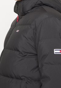 Doudoune noire avec fermeture éclair sur le devant, col haut et tirette rouge. Présente un patch logo sur la manche et une texture matelassée.