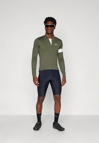 Cyklistický outfit zahrnující tmavozelenou dlouhý rukáv dres, s předním zipem, bílými akcenty a fitted černé šortky. Černé cyklistické boty a sluneční brýle.