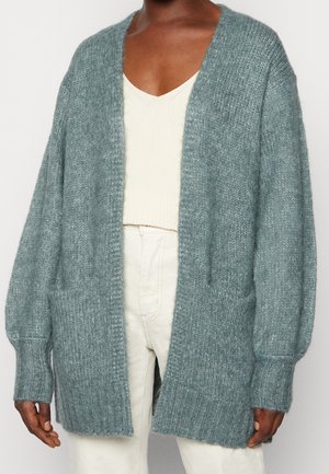 Person wearing a loose, soft grayish-blue knitted cardigan over a white top and cream-colored pants against a plain background.