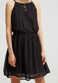 Robe en chiffon noire avec un corsage ajusté, des accents de dentelle sur les épaules et une taille élastique. Jupe fluide avec une légère brillance.