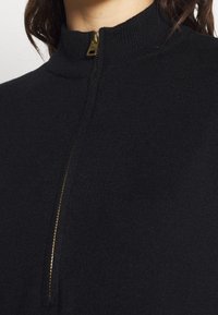 Pull noir en tricot avec un col montant et des détails de fermeture éclair dorée au niveau du col, présentant une texture lisse et un design simple.