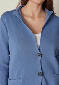 Blå, stickad cardigan med V-hals, två framfickor och två mörka knappar. Mjuk textur och avslappnad passform.