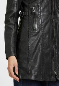 Giacca in pelle nera con zip frontale, tasca laterale e vita con cintura. Presenta una texture liscia e dettagli di cucitura ben visibili.