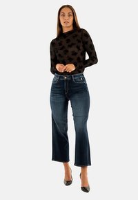 Zwarte top met lange mouwen en bloemenpatroon, gecombineerd met blauwe denim cropped wide-leg jeans. Zwarte hoge hakken maken de outfit compleet.