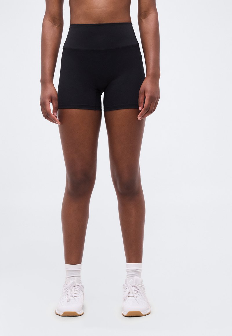 Άτομο που φοράει μαύρα αθλητικά σορτς με ψηλή μέση, λευκές κάλτσες και λευκά αθλητικά παπούτσια, στέκεται μπροστά σε απλό ανοιχτό φόντο.