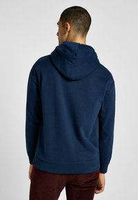 Sudadera azul marino con una textura suave, capucha y puños y dobladillo acanalados. La vista trasera muestra un diseño simple y estilizado sin adornos.