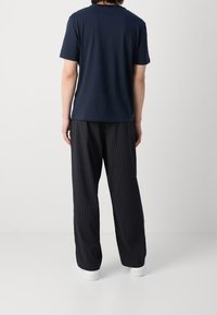 T-shirt di cotone blu navale a maniche corte, abbinato a pantaloni neri a righe verticali e sneakers bianche, visto da dietro su uno sfondo neutro.