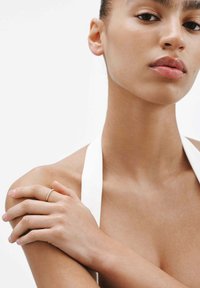 Joven mujer con piel suave, usando un anillo delgado en su mano izquierda, descansando la mano sobre su hombro derecho, vestida con un top halter blanco.