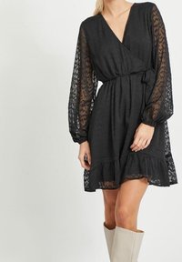 Svart omlottklänning med långa genomskinliga ärmar, texturerad tyg, midja med bälte och rynkad fåll. Accenterad med ett spetsmönster.