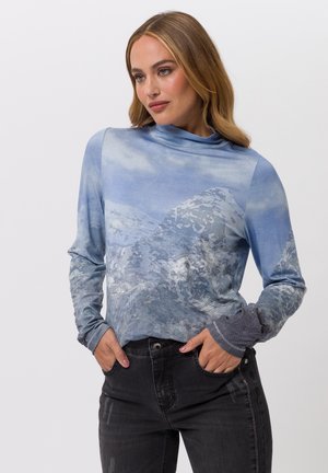 Woman with long blonde hair wearing a blue long-sleeve top with mountain print and black jeans, hands in pockets, looking right.