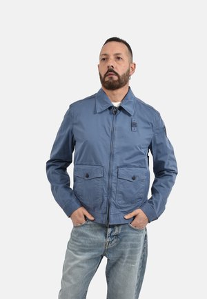 Uomo con barba che indossa una giacca blu con cerniera e jeans azzurri chiari, in piedi con le mani nelle tasche su uno sfondo bianco uniforme.