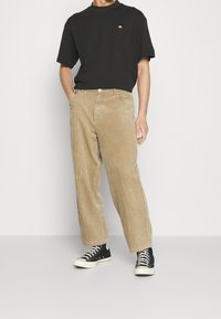 T-shirt noir à manches courtes avec un petit logo rouge et orange, associé à un pantalon en velours côtelé beige et des baskets montantes noires.