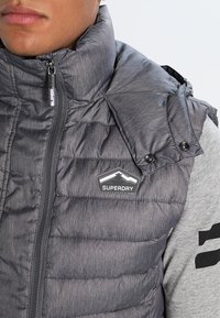 Grå pufferväst med hög krage, kviltad textur och svarta detaljer. Har en Superdry-logotyp på framsidan och knapppynt.