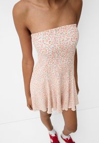 Axellös, blommig klänning i ljusrosa med små gröna och orange blommor. Utsvängd kjol med vågig fåll. Modell som bär röda sneakers.