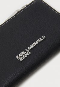 Svart läderplånbok med en texturerad yta, prydd med vit sömnad och en silvrig "KARL LAGERFELD JEANS"-logotyp. Dragkedjestängning.