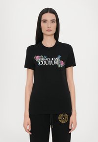 Juodas trumpaapykaklis marškinėliai, puošiami gėlių siuvinėjimu ir baltu "VERSACE JEANS COUTURE" logotipu. Minkšta medvilnė. Standartinė fasonas.