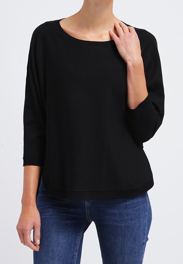 Femme portant un haut noir ample à manches trois-quarts et un jean bleu, avec une main touchant son cou et l'autre le long du corps.