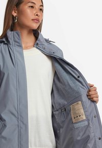 Ljust grå vattentät jacka med silverdetaljer, huva och innersidetikett. Slät yttertextur och mjukt foder synligt.