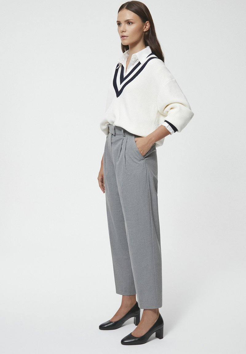 Biały sweter dzianinowy z granatowymi paskami przy dekolcie w kształcie V, w połączeniu z szarymi spodniami z wysokim stanem i plisami oraz czarnymi butami na obcasie.