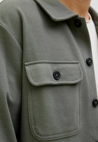 Olive-green jacket with a textured finish, featuring a pointed collar, black buttons, and a large chest pocket.