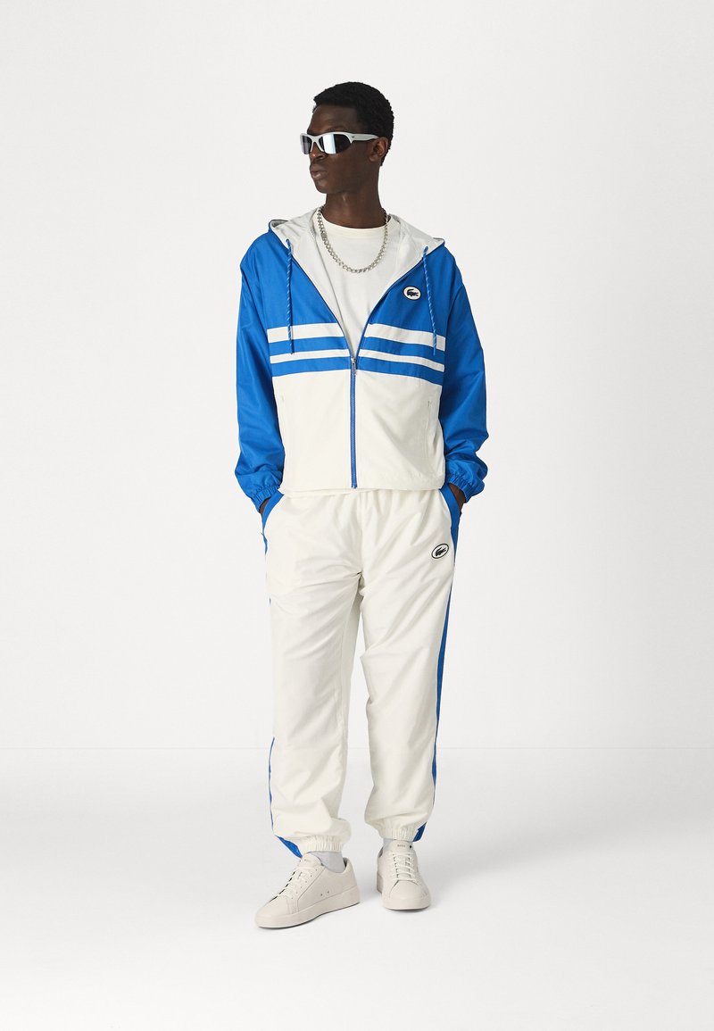 Tuta blu e bianca con giacca zip-up, due strisce orizzontali, polsini elasticizzati e pantaloni coordinati, abbinata a sneaker bianche e occhiali da sole.