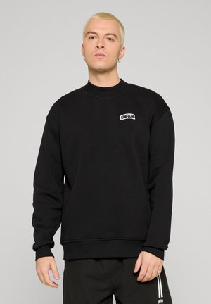 Una giovane persona con capelli biondi corti indossa una felpa nera con il logo "UNFAIR" sul petto, in piedi davanti a uno sfondo grigio chiaro.