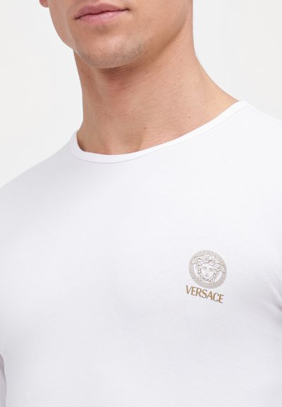 Weisses Baumwoll-T-Shirt mit rundem Ausschnitt und kurzen Ärmeln, verziert mit einem goldenen, gestickten Versace-Logo auf der Brust.
