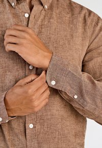 Bruin linnen overhemd met knopen, gemaakt van een gestructureerde stof, met witte knopen en verstelbare manchetten. Handen raken de stof van het overhemd aan.