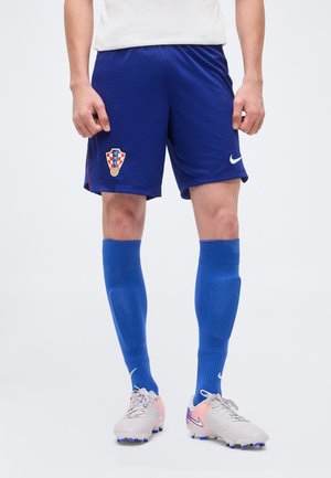 Muž oblečený v modrých futbalových šortkách Chorvátska, s rovnakými modrými ponožkami a bielymi futbalovými kopačkami Nike s ružovými akcentmi, stojaci na bielom pozadí.