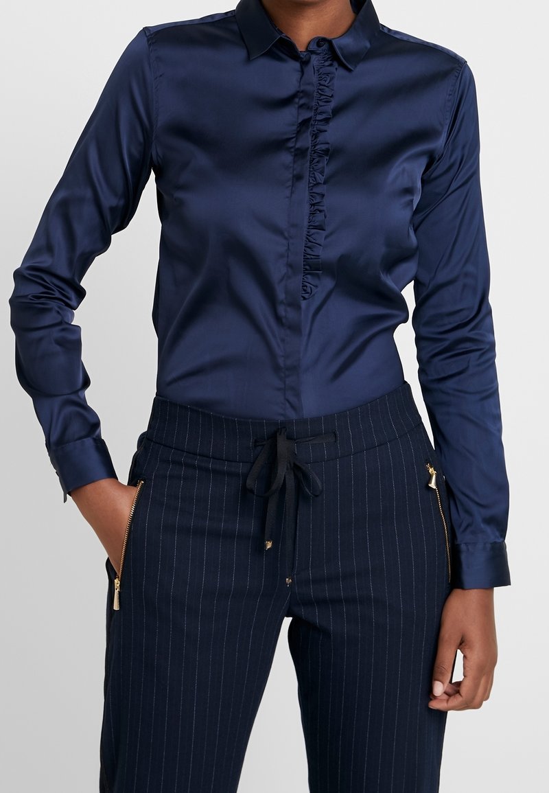 Femme portant un chemisier en satin bleu foncé avec détails volantés et un pantalon à fines rayures bleu marine avec poches zippées et taille cintrée à cordon.