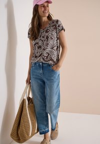 Femme portant une casquette rose, une chemise marron à imprimé cachemire, un jean bleu, tenant un grand sac cabas tissé et souriant sur un fond beige.