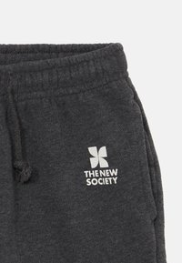 Tumši pelēki sporta bikses ar aukliņu jostu un baltu logo, kas lasa "JAUNĀ SOCIETĀTE" ar tauriņa dizainu.