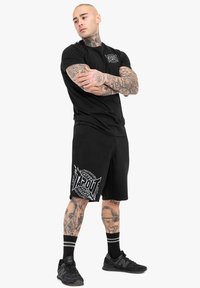 Černé tričko s krátkým rukávem a logem, spárované s černými kraťasy s bílou grafikou TAPOUT. Na nohou má černé sportovní boty a pruhované ponožky.