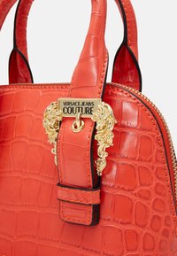 Červená kabelka s krokodýlí strukturou, dvojitými uchy a zlatou ozdobnou destičkou s logem "Versace Jeans Couture" na popruhu.