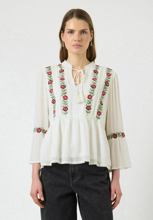 Moteris dėvi baltą palaidinę su gėlių siuvinėjimu, varpelio formos rankovėmis ir kaklo raišteliu, derinančią su juodais džinsais ant vientisos foninės.