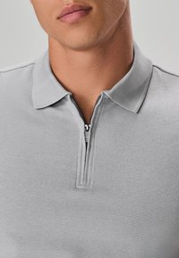 Graues Poloshirt aus strukturiertem Stoff, mit kurzem Kragen und einem Reißverschluss am Ausschnitt. Der Reißverschluss hat einen metallischen Zuggriff.