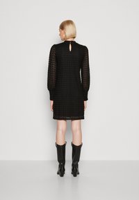 Vestido preto com padrão texturizado, mangas longas transparentes e detalhe das costas em forma de chave. Combinado com botas pretas até ao joelho.