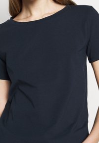 Kobieta w prostym, ciemnoniebieskim T-shircie z krótkim rękawem o okrągłym dekolcie, widoczna od ramion do bioder.