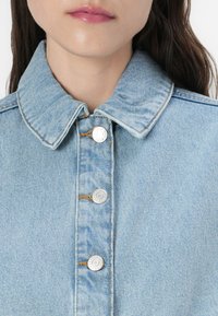 Casaco de ganga azul claro com colarinho clássico, botões prateados e costura laranja em contraste ao longo da frente e das costuras.