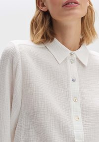 Camicia bianca con texture, caratterizzata da un colletto classico, una pattina con bottoni perlacei e un motivo di tessuto stropicciato.