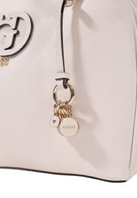 Borsa bianca con texture e rifiniture nere, dotata di un anello charme dorato, inclusi un'etichetta circolare "GUESS" e un piccolo ciondolo decorativo.