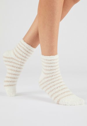 Piernas con calcetines blancos y peludos con rayas horizontales beige claro sobre un fondo claro y liso.
