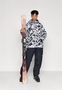 Inimene mustade suusapükste ja saapatega, kes hoiab vertikaalselt suuski, kätel on mustad suusaprillid.