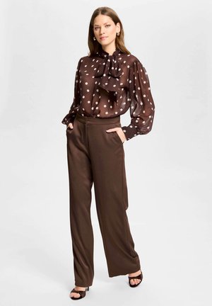 Staande vrouw met een doorzichtige bruine blouse met polkadots en een das, brede bruine broek en zwarte open-teenhakken, handen in de zakken.