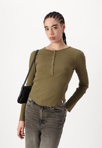 Oliven grønn ribbet langermet topp med rund hals og fem knapps placket, kombinert med lysegrå denimbukser. Svart håndveske som aksent.